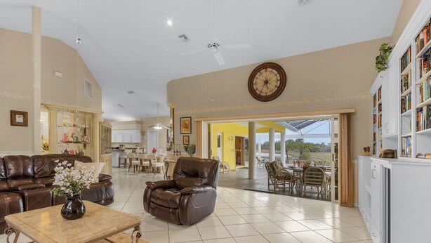 A spacious living room with a leather sofa, a recliner, and a coffee table. There is a large clock on the wall and a bookshelf filled with books. - The Yellow | Lakefront Home, Heated Pool & Spa (Leigh Acres)