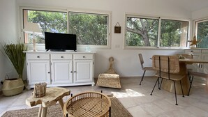 Ein Zimmer mit Fernseher auf einer weißen Kommode, Korbmöbeln und großen Fenstern mit Blick auf Bäume.