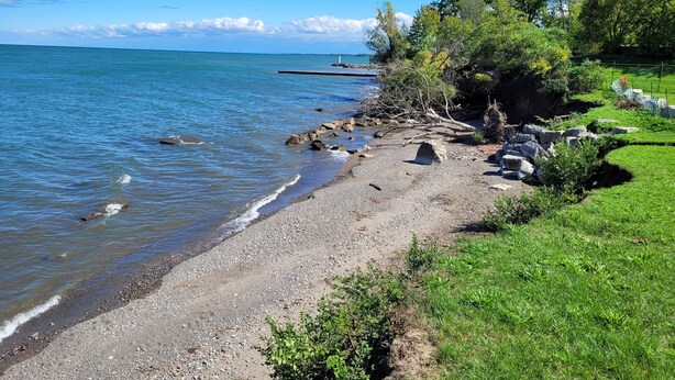 Une plage de galets avec une zone herbeuse et des arbres, un phare visible au loin, et une eau calme.