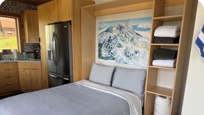 Quarto com cama, pintura de uma montanha, frigorífico e prateleira com toalhas dobradas.