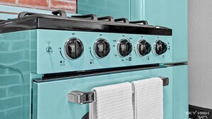 A teal-colored stove with four knobs and a white towel hanging on a rack.
