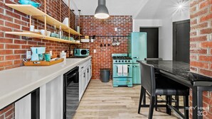 A compact kitchen with a brick wall, wooden shelves, a teal refrigerator, and a black dining table.