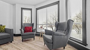A modern living room with grey armchairs, a red pillow, and large windows with blinds.