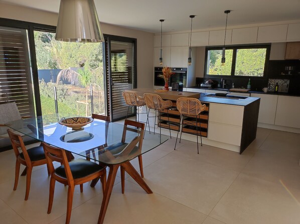 A modern kitchen with a glass dining table, wooden chairs, and a central island. - Contemporary Villa with Ocean Views (Hyères)