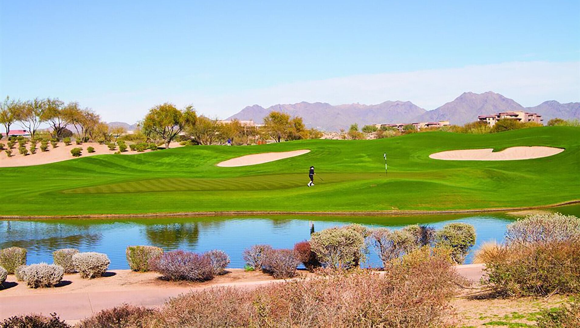 Campo de golf con un estanque, búnkeres de arena y montañas al fondo.