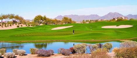 Campo de golf con un estanque, búnkeres de arena y montañas al fondo.