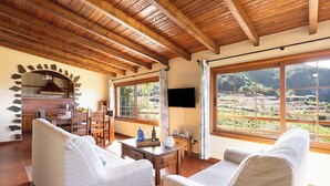 Un salon chaleureux avec des poutres en bois, une cheminée en pierre, un téléviseur et de grandes fenêtres donnant sur un paysage.