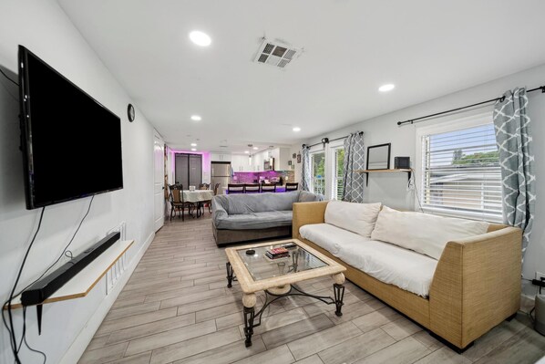 A modern living room with a flat-screen TV, a sofa, a coffee table, and a kitchen area in the background.