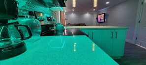 A modern kitchen with a teal countertop, a coffee maker, and a kettle. There is a TV mounted on the wall and a seating area with a sofa and cushions.