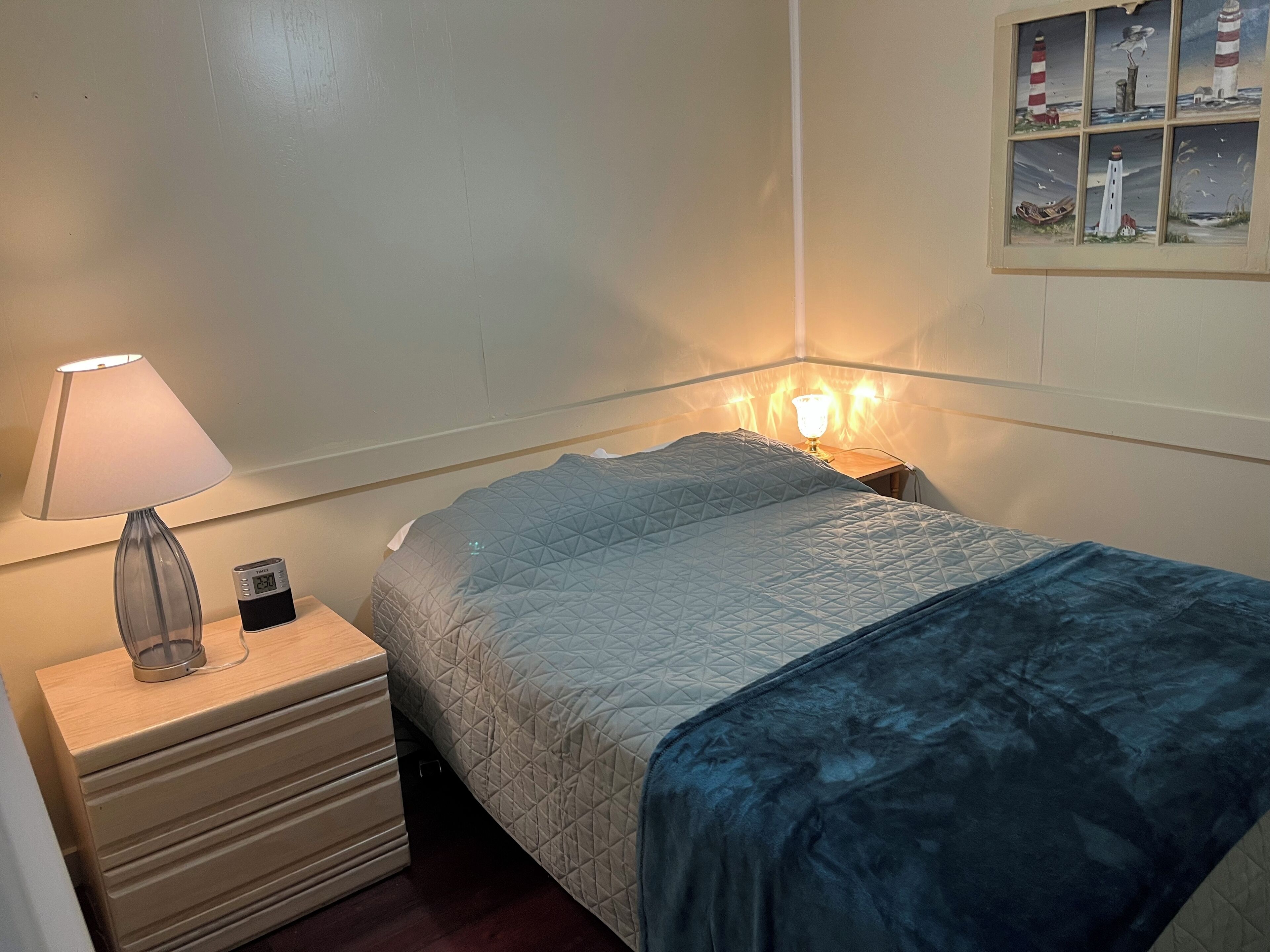 A neatly made bed with a quilted blue blanket, a bedside lamp, and a small bedside table with a clock.