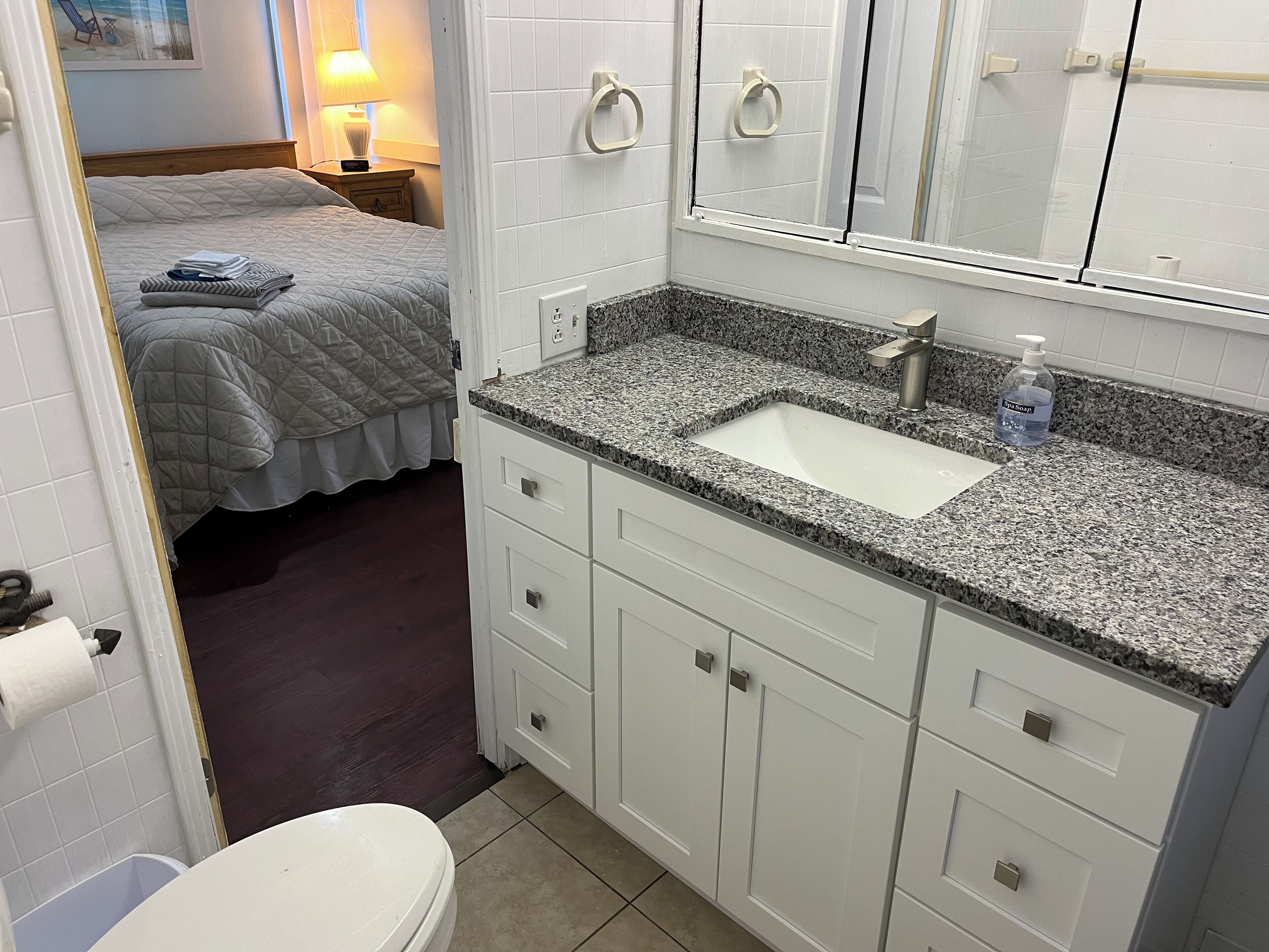 Un baño con lavabo, espejo e inodoro. Se observa una cama con manta acolchada y una mesita de noche con lámpara.