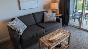 A dark grey sofa with patterned cushions, a wooden coffee table with a tray, a lamp, and a painting of a landscape on the wall. - Beachfront Walk-out in the Heart (Qualicum Beach)