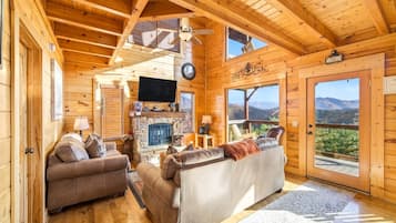 A log cabin interior with a fireplace, two sofas, a television, and a scenic mountain view.
