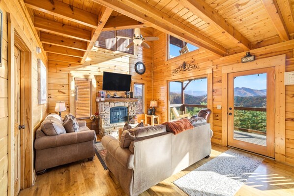 A log cabin interior with a fireplace, two sofas, a television, and a scenic mountain view.