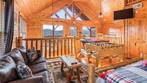 Ein gemütliches Holzhaus mit Schlafzimmer im oberen Bereich, einem Wohnbereich mit Sofa und Kicker sowie einem Balkon mit Aussicht.
