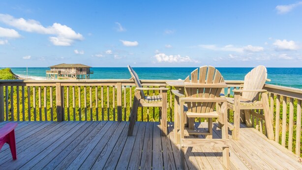 Eine Holzterrasse mit Stühlen und einem Tisch mit Blick auf einen Strand und ein Gebäude mit einem Reetdach.