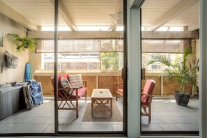 Une terrasse ensoleillée avec une table et des chaises en bois, un pot de fleurs et une vue sur les bâtiments à travers de grandes fenêtres.