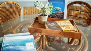A round glass table with a wooden tray, a potted plant, and books. There are two wicker chairs around the table. - Oceanfront Condo/Hawaiian Style. 1Bd, w/AC & Wifi (Kailua-Kona)