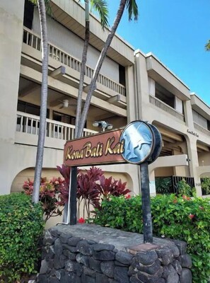 Une enseigne pour Kona Bali Kai, avec une base en pierre et un emblème circulaire, entourée de verdure et de palmiers.
