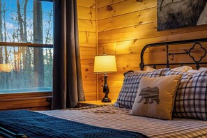 Ein gemütliches Holzblockhaus-Zimmer mit einem Bett, einem Nachttisch mit Lampe und einem Fenster mit Vorhängen.