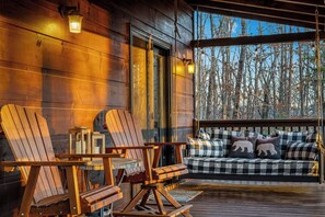 Eine Holzterrasse mit Adirondack-Stühlen und einem karierten Sofa mit Bärenkissen, mit Blick auf einen Wald.