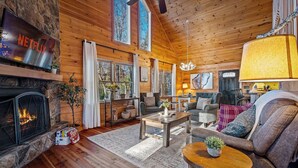 A cozy log cabin interior with a fireplace, a Netflix sign, a chandelier, and a large window overlooking a forest. - Private Hiking Trail, Full Game Rm, Great Location (Morganton)
