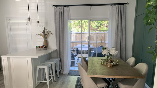 A dining area with a wooden table, white chairs, a patterned rug, and a view through a glass door to an outdoor patio.