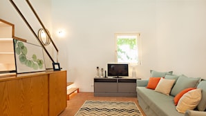 Una sala de estar con un aparador de madera, un televisor sobre un mueble gris, un sofá verde con cojines naranja y blancos, y una alfombra estampada.
