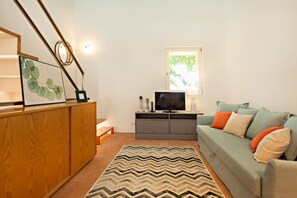A living room with a wooden cabinet, a television on a grey stand, a green sofa with orange and white cushions, and a patterned rug.