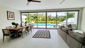 A spacious living area with a dining table, sofas, and a large sliding glass door leading to a pool. - Mama Taras Luxury Villa (Rarotonga)