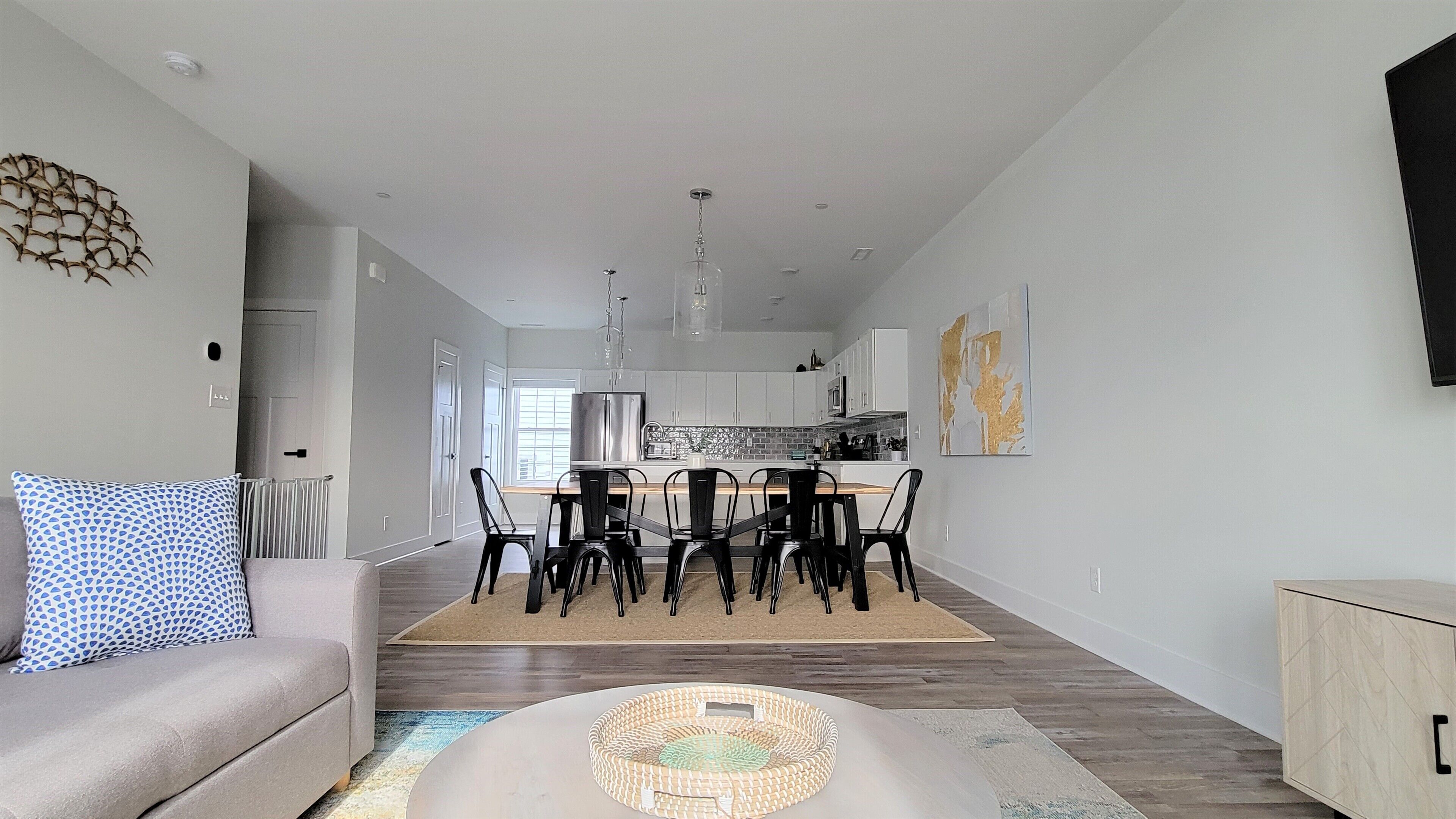 Una sala de estar moderna con sofá, mesa de comedor y sillas. Se observa una cocina al fondo con gabinetes blancos y un refrigerador.