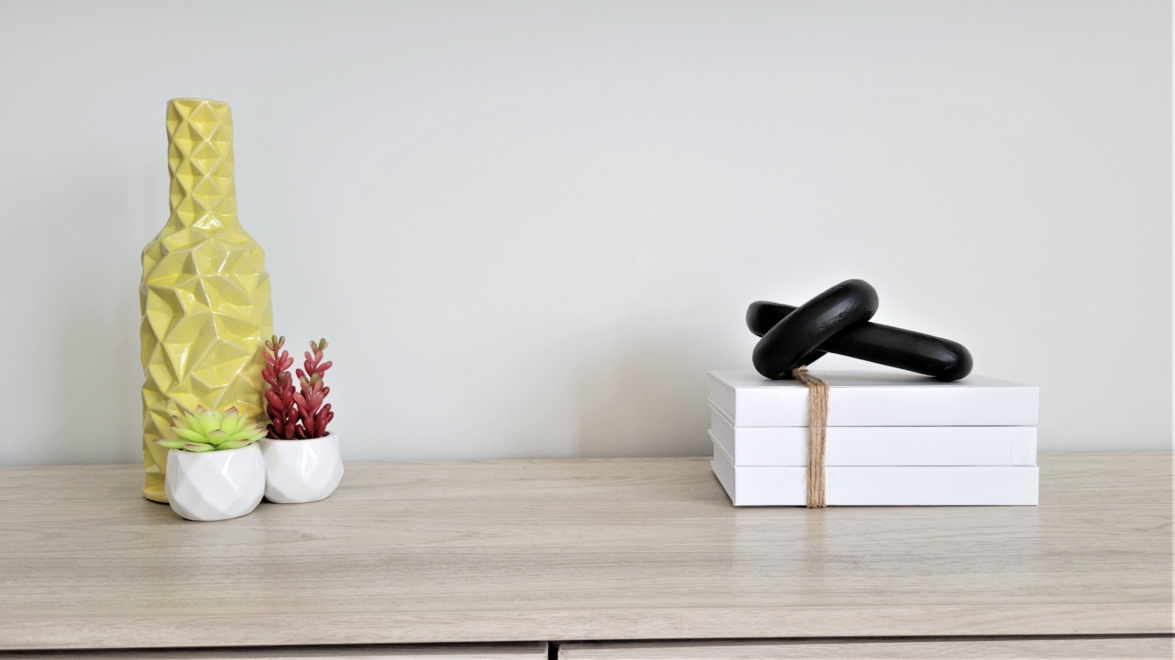 A wooden shelf with a yellow vase, two small potted plants, and a stack of white boxes with a black object on top.
