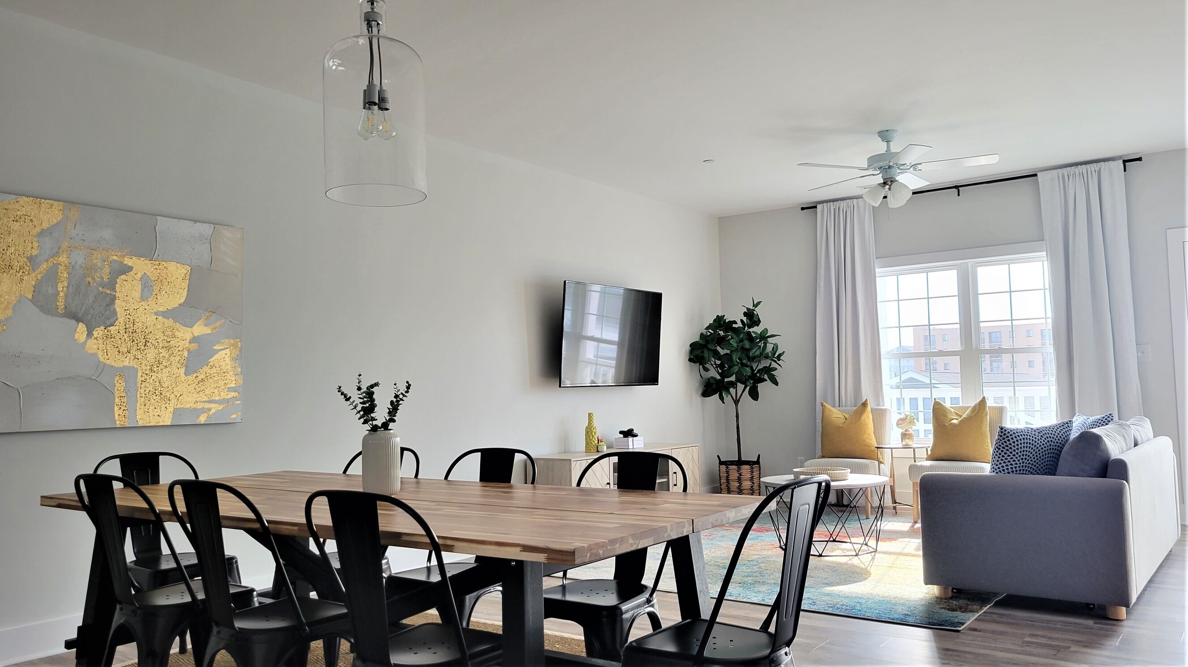 A modern living room with a dining area, featuring a wooden table, black chairs, a grey sofa, a ceiling fan, and a large window with curtains.