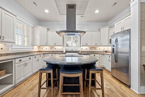 Una cocina moderna con armarios blancos, una isla central con taburetes, un refrigerador de acero inoxidable y un ventanal con persianas.
