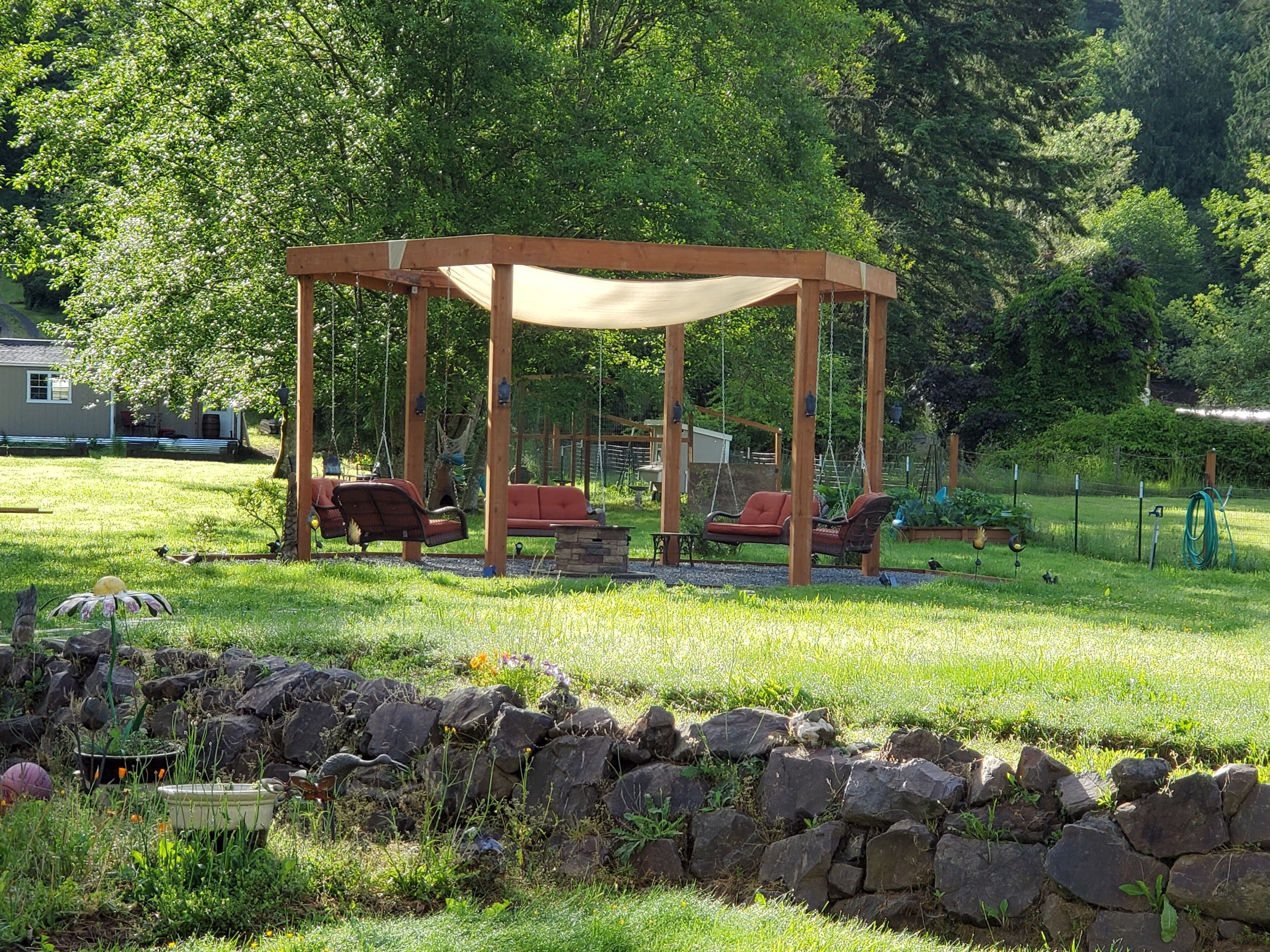 Un gazebo in legno con tettoia, circondato da un muro di pietra, in un prato erboso con alberi e una casa sullo sfondo.
