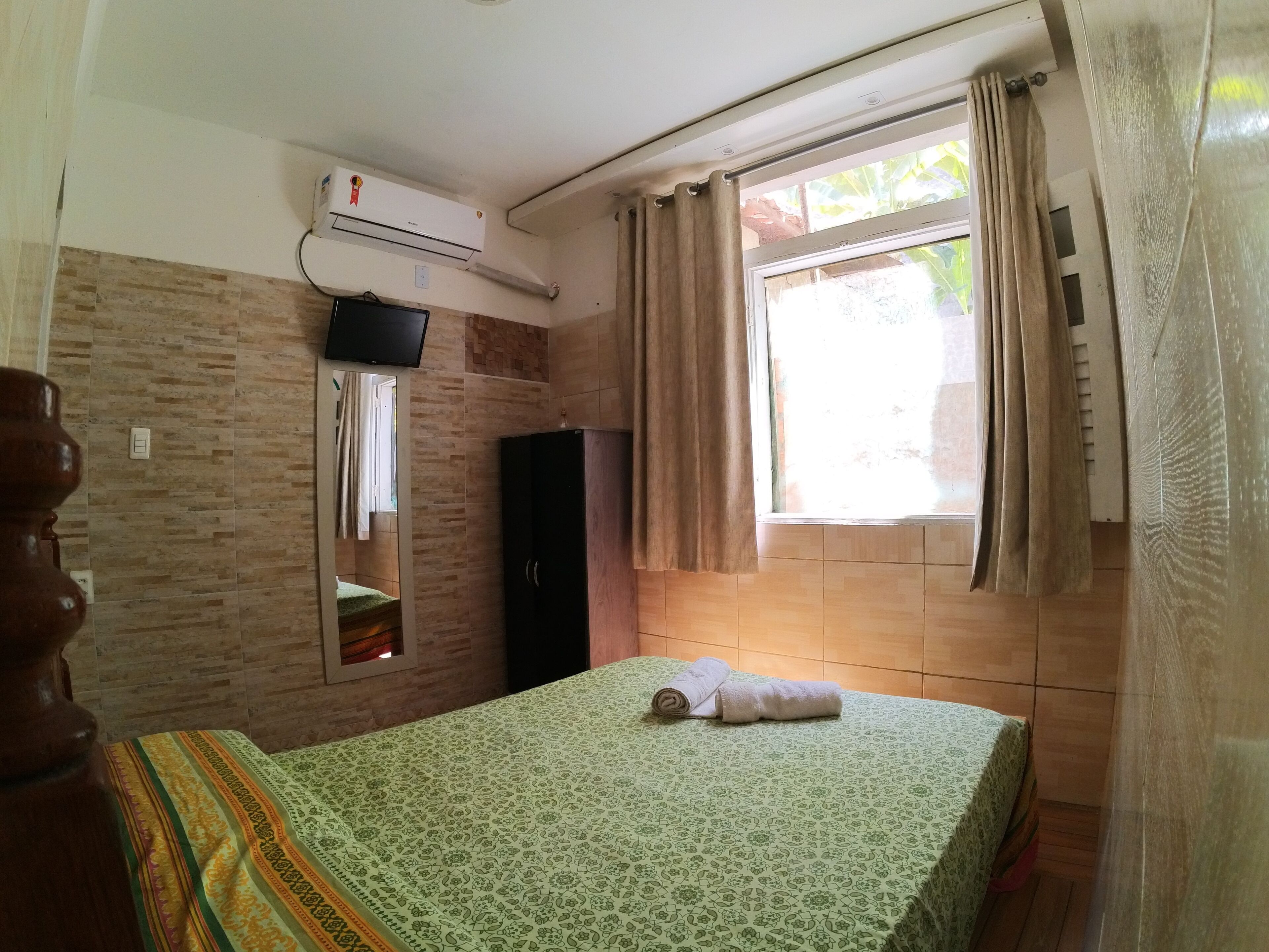 Quarto com cama, guarda-roupa, televisão na parede e janela com cortinas.