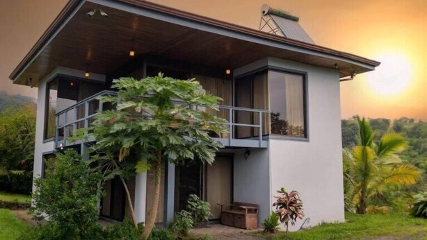 Una casa moderna de dos pisos con balcón, rodeada de abundante vegetación.