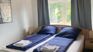 Un lit double avec une literie bleue, une fenêtre avec des rideaux, une petite table de chevet et un tableau encadré au mur.