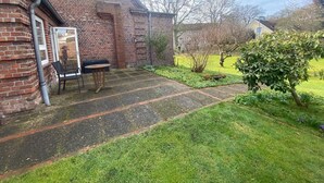 Un bâtiment en briques avec une fenêtre, une porte et un petit espace jardin agrémenté d’un banc et d’une table.