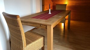 Une table de salle à manger en bois, surmontée d’un set de table rouge, avec deux chaises en osier et une bougie.