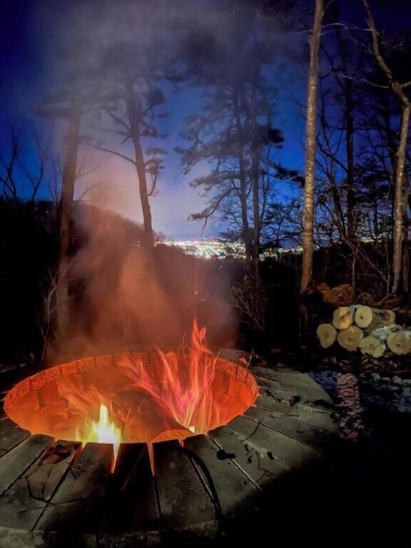 Un feu de camp dans une zone boisée, entouré d’arbres, avec une vue lointaine sur une ville.