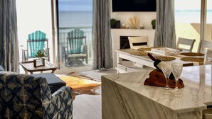 Un salón moderno con un televisor grande, un comedor con una mesa dispuesta para dos personas y vistas al océano a través de amplios ventanales.