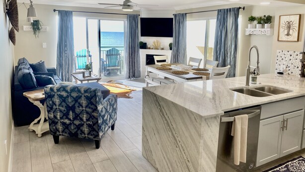 Una sala moderna con un sofá, un sillón estampado, una mesa de comedor con individuales y una cocina con encimera de mármol y electrodomésticos de acero inoxidable.
