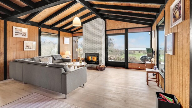 A modern living room with a grey sectional sofa, a fireplace, wooden beams, and large windows offering a view of the outdoors.