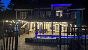 Una casa de dos pisos con piscina, iluminación exterior y un patio cubierto.