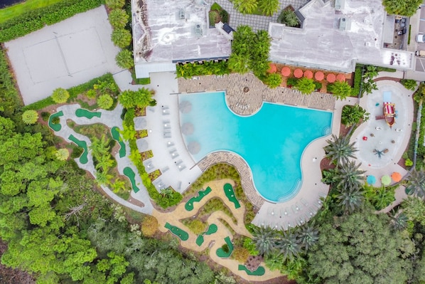 An aerial view of a resort with a large swimming pool, multiple lounge areas, and a golf course.