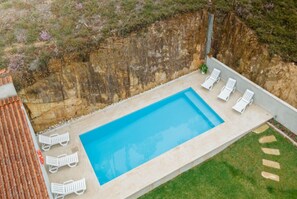 A rectangular swimming pool with a clear blue surface, surrounded by a low stone wall. Adjacent to the pool, there are several white lounge chairs aligned in a row.