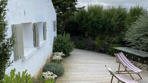 A wooden deck with two lounge chairs and a hammock, surrounded by lush greenery and a white building with shutters.