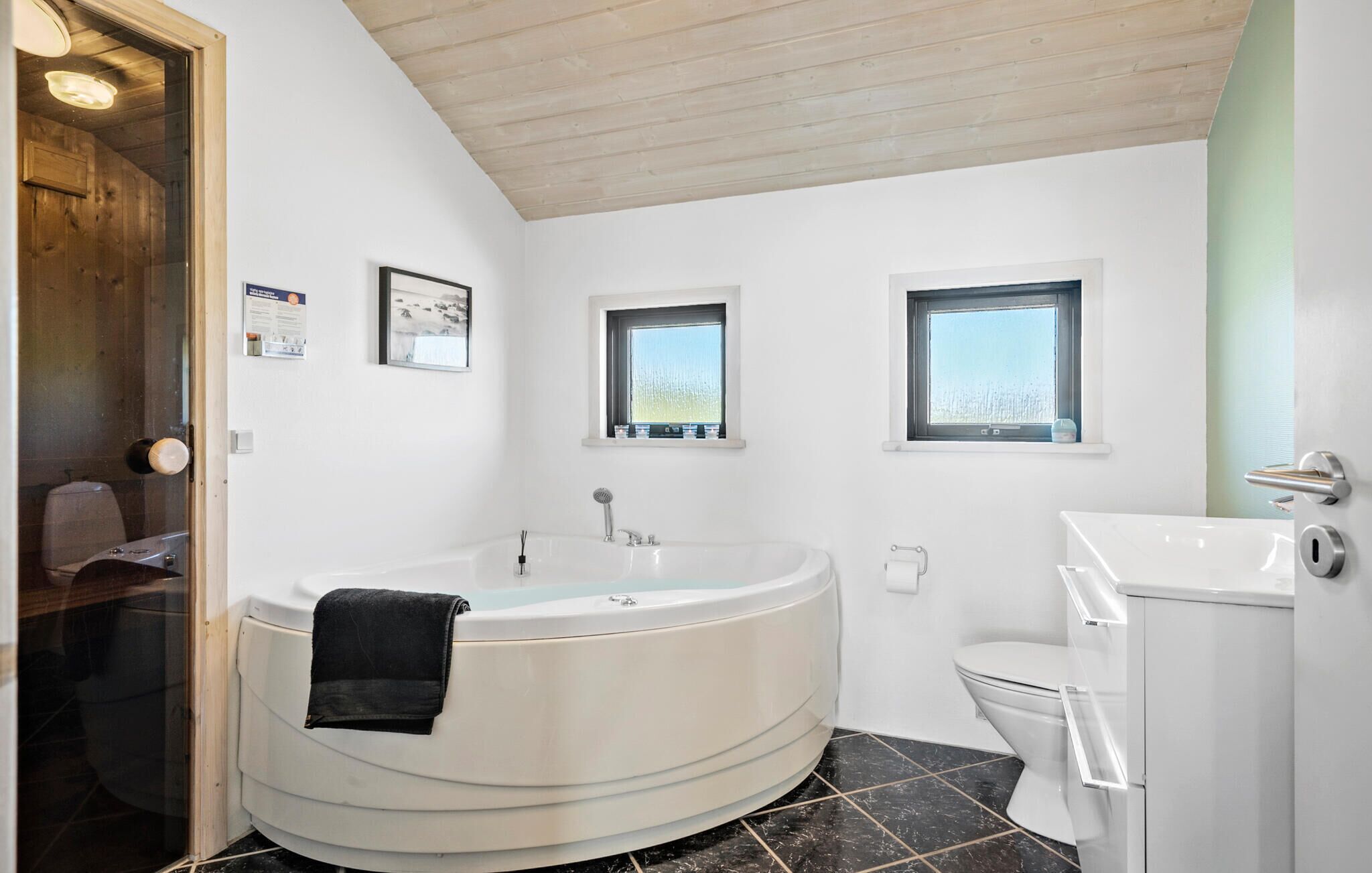 A modern bathroom with a freestanding bathtub, a toilet, and a sink. There are two windows providing natural light.