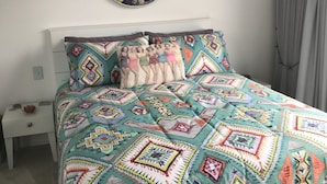 Une chambre avec une couvre-lit colorée à motifs matelassés, un élément décoratif rond fixé au mur et une table de chevet blanche surmontée d’une petite lampe.